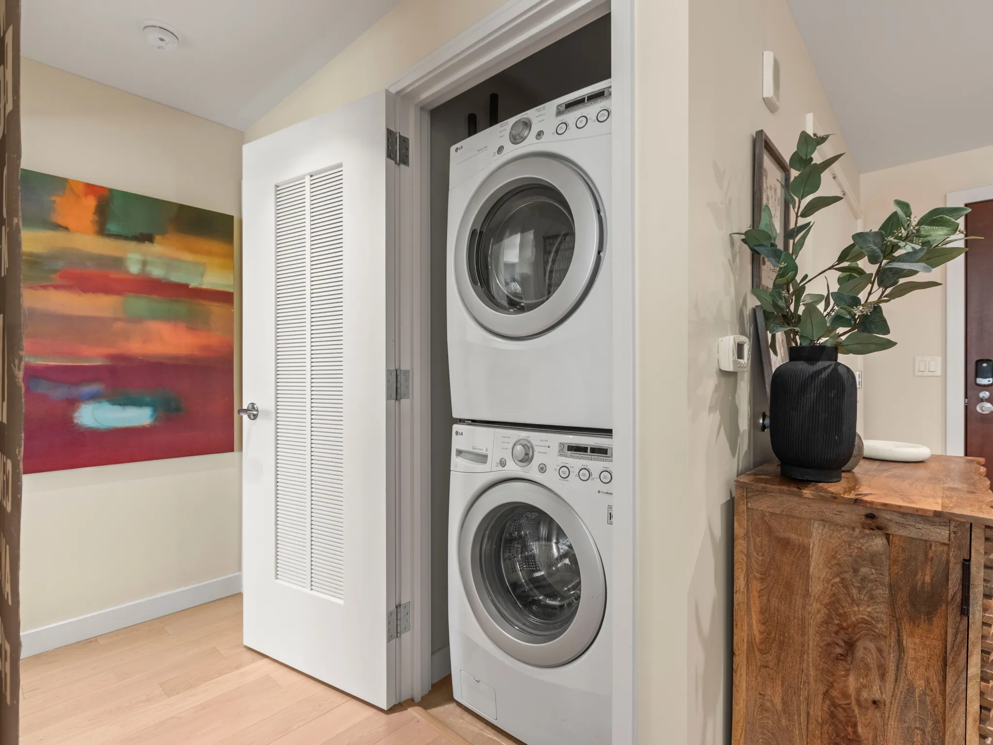 Integrated laundry closet with stacked washer and dryer, light wood-finish flooring, and a louvered door