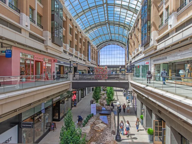 Multi-level retail concourse with a glass-paneled arched ceiling, featuring a central water feature with natural rock elements, multiple storefronts, and elevated walkways with glass railings