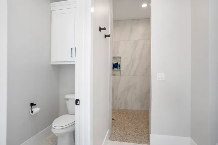 White built-in cabinetry, white toilet, shower with large format tile, shower with stone flooring, and recessed shower niche