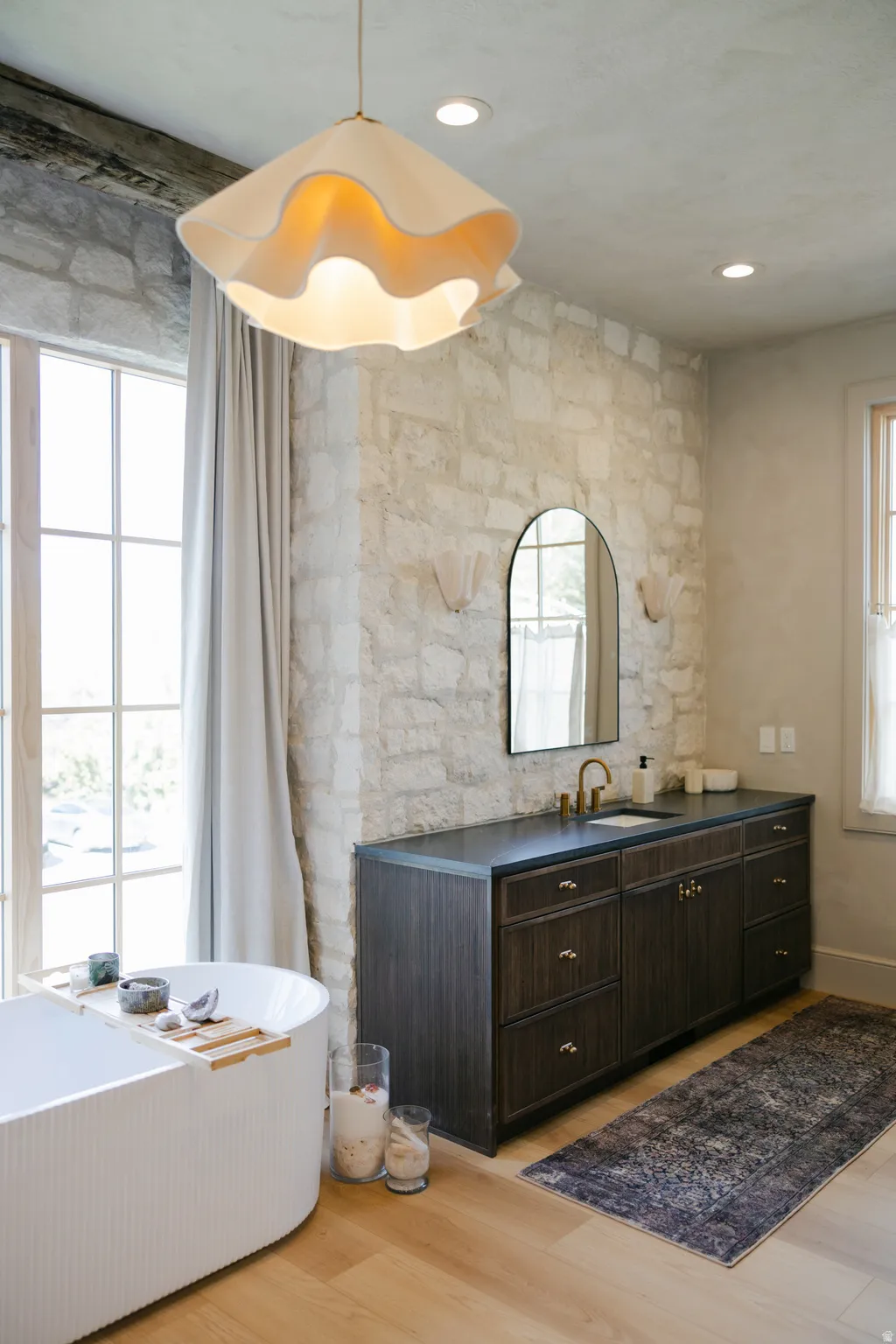Freestanding bathtub with ribbed exterior, stone accent wall, wood-finish flooring, dark wood vanity with integrated sink, and an arched mirror