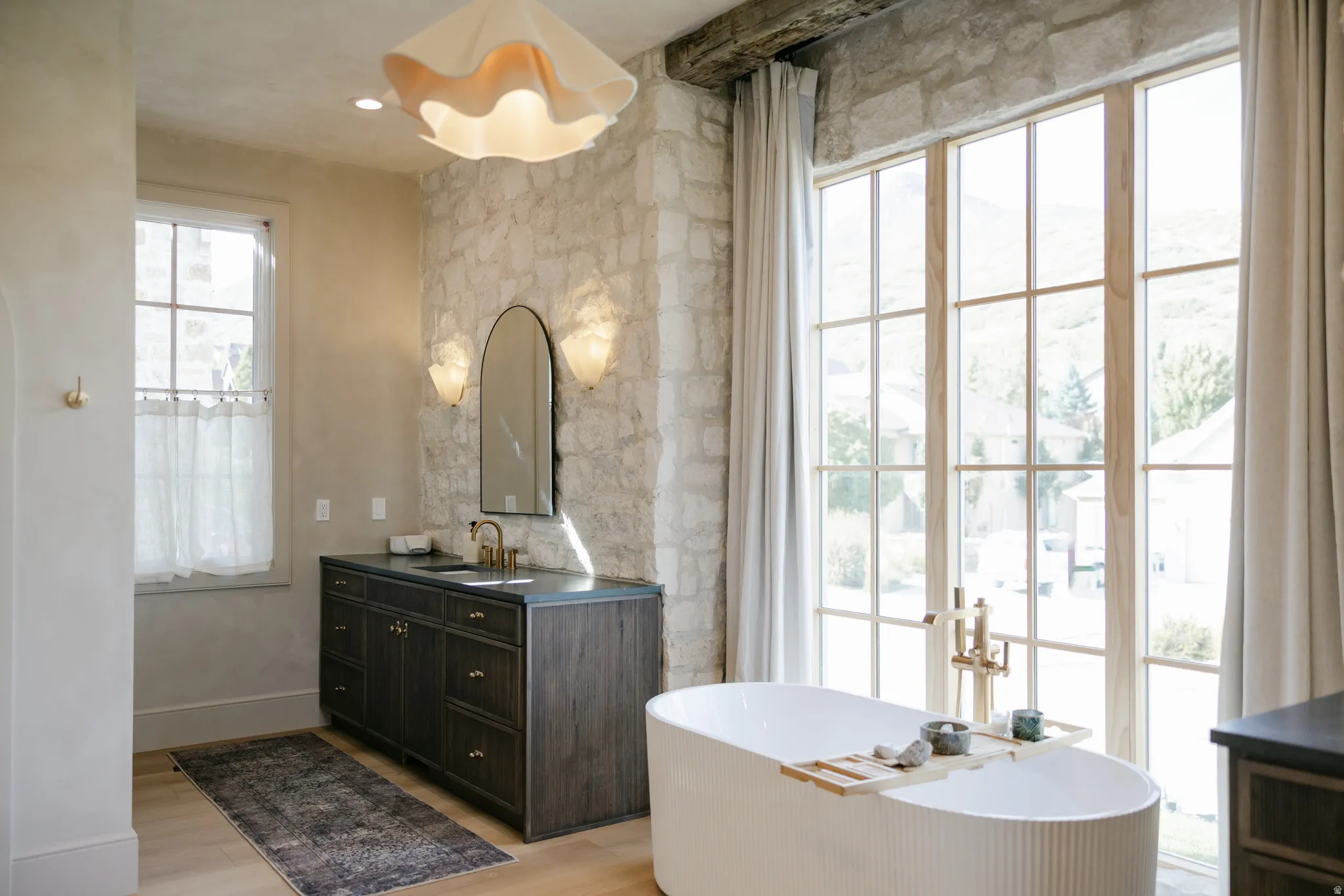 Stone accent wall, floor-to-ceiling windows, freestanding soaking tub, single vanity with dark cabinetry, and light wood-finish flooring