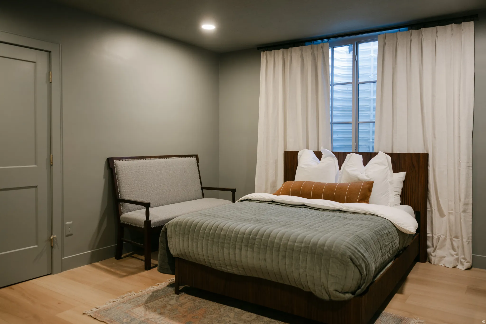 Recessed lighting illuminates a room featuring light wood-finish flooring, a solid panel door with brass-tone hardware, and a window with white drapes