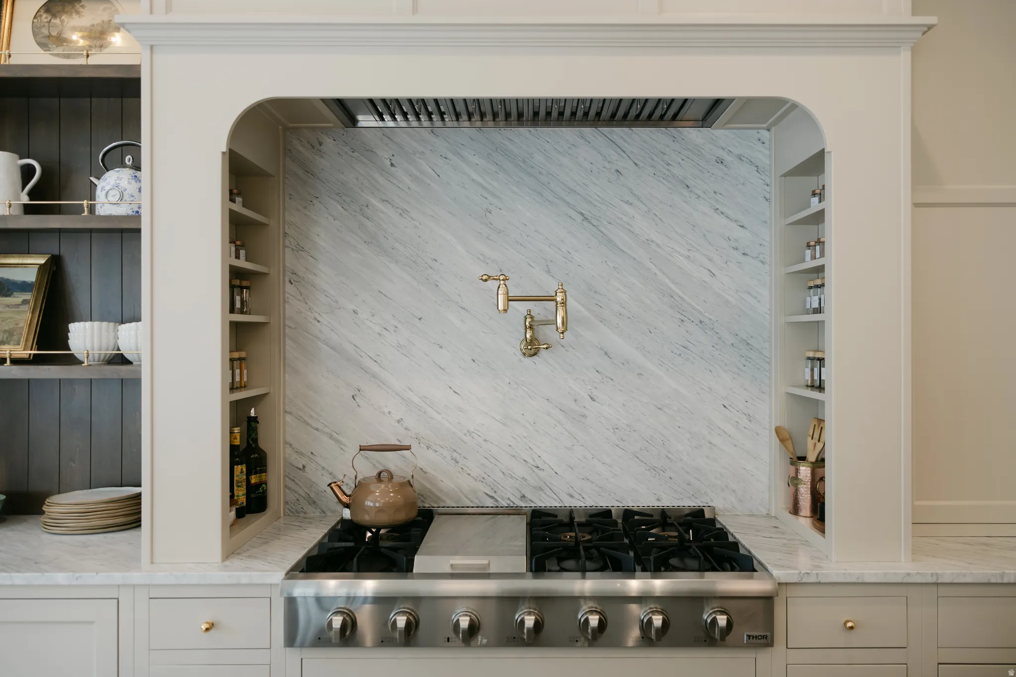Stainless steel gas range with a pot filler, integrated range hood, marble slab backsplash, built-in shelving, and light-toned cabinetry