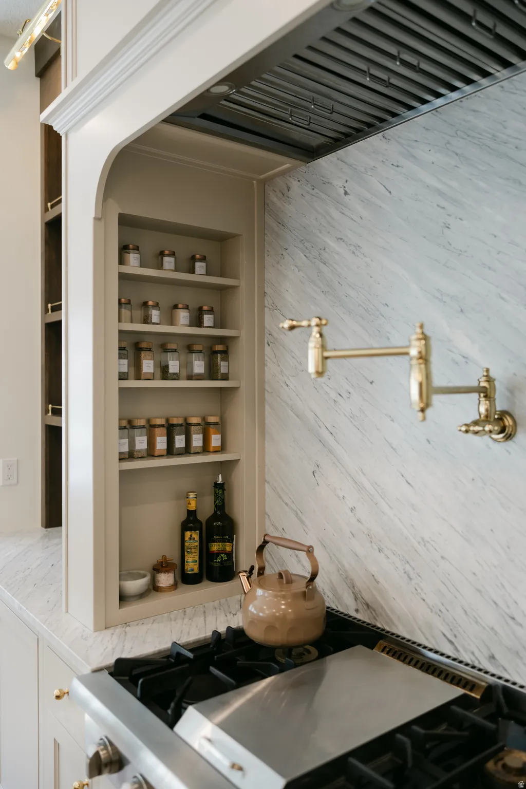 Built-in spice rack, polished brass pot filler, stainless steel range with griddle, natural stone backsplash, and matching natural stone countertop