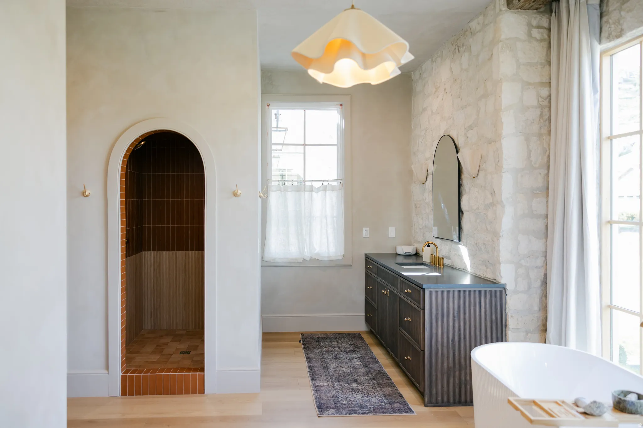 Arched shower entry with terra cotta tile, stone accent wall, double vanity with dark wood finish, and freestanding bathtub