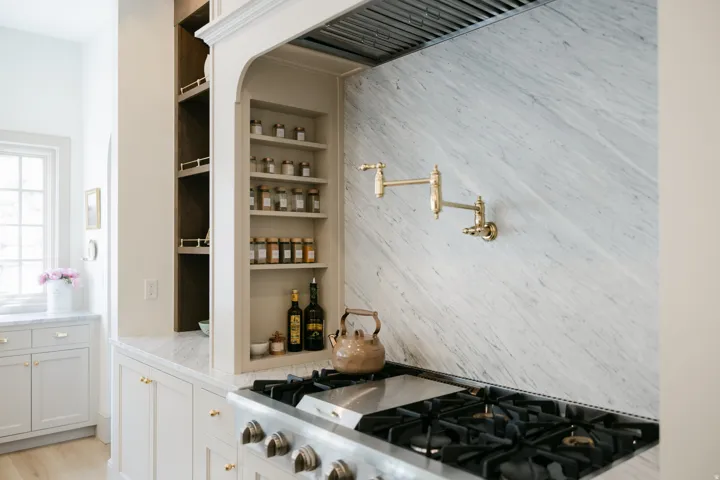 Built-in gas range, stone slab backsplash, wall-mounted pot filler, custom cabinetry with open shelving, and light-toned countertops