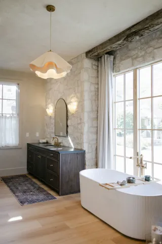 Freestanding soaking tub with fluted detailing, wide plank wood-finish flooring, stone accent wall, large picture windows, and a dark wood vanity with a bronze faucet
