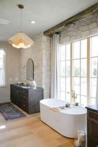 Freestanding soaking tub with floor-mounted faucet, wood-finish flooring, stone accent wall, large window with grid pattern, and a dual vanity with undermount sinks