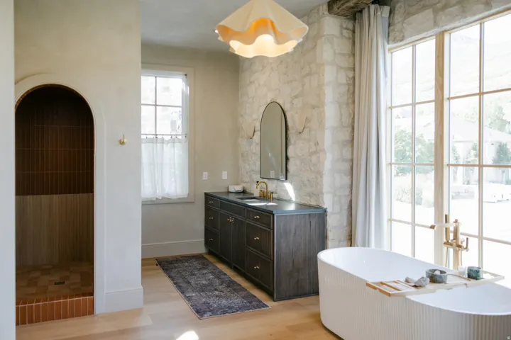 Freestanding fluted soaking tub, arched walk-in shower with vertical tile, wood-finish flooring, stone accent wall, and a dark wood-finish vanity with dual sinks