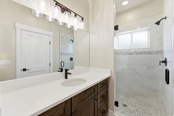 Dark wood-finish vanity with white countertop, matte black faucet, and clear glass shower enclosure featuring a mosaic tile accent and pebbled shower floor