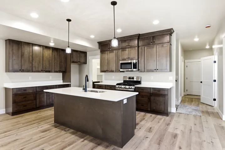 Kitchen featuring wood-finish flooring, dark wood cabinetry, white countertops, a central island with an integrated sink, and stainless steel appliances