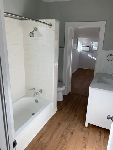 Bathtub with white subway tile surround, wood-finish flooring, white vanity with integrated sink, and white toilet