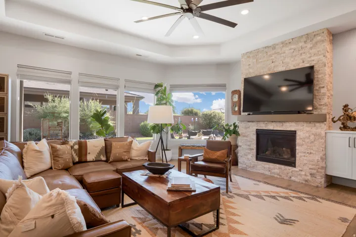Tray ceiling with a ceiling fan, stone-clad fireplace with a wood mantel, built-in cabinetry, and multiple windows