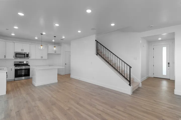 Open-concept living space featuring wood-finish flooring, a contemporary kitchen with white cabinetry and stainless steel appliances, and a staircase with a black metal railing