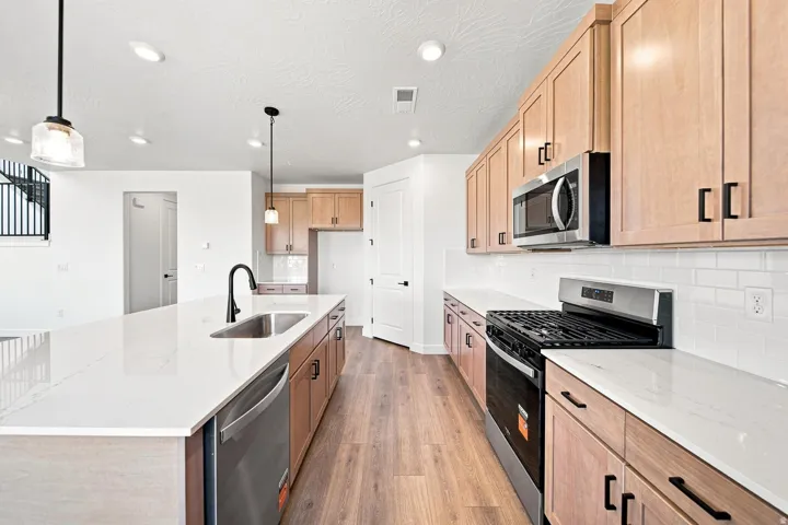 Kitchen featuring a large island with an undermount sink, stainless steel appliances, light wood-finish cabinetry, white subway tile backsplash, and wood-finish flooring