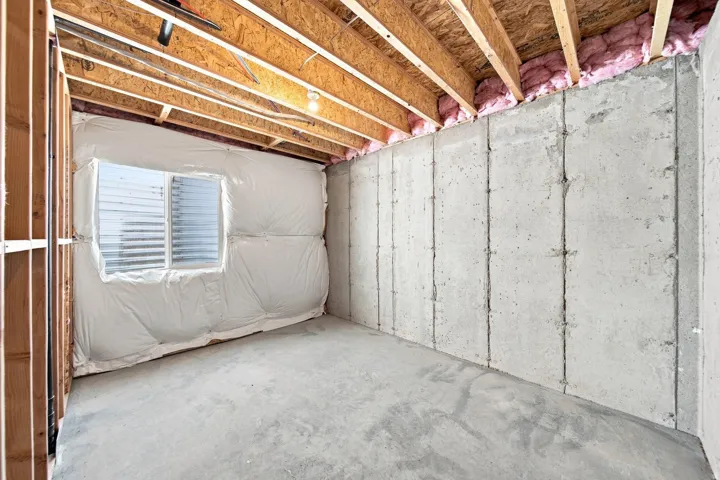 Unfinished basement space with exposed wooden ceiling joists, concrete flooring, concrete foundation walls, a single window, and visible insulation