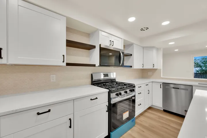 White shaker cabinetry with matte black hardware, stainless steel appliances, light-toned tile backsplash, solid surface countertops, and wood-finish flooring