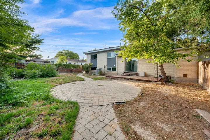 Expansive backyard featuring a paver patio and walkway, mature trees, and a ground-level wood deck with sliding glass door access