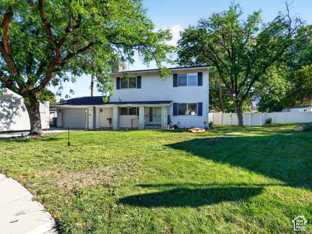 Two-story exterior with white siding, dark shutters, an attached garage, a front lawn, and mature trees
