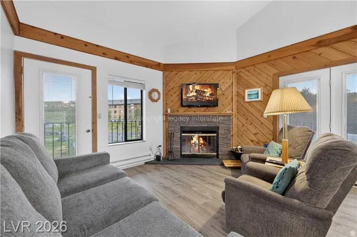 Vaulted ceiling, wood-finish flooring, wood-paneled accent wall, stone-front fireplace, and balcony access