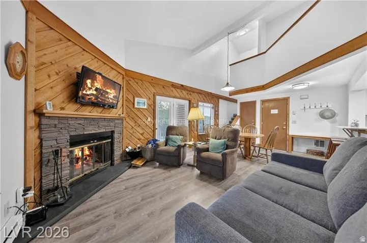 Vaulted ceilings with exposed beams, stone-front fireplace, wood paneling, sliding glass door, and wood-finish flooring