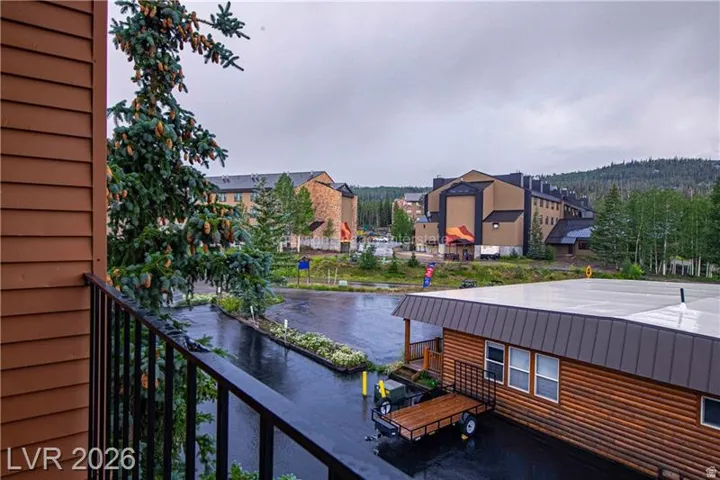 Brown horizontal siding with a black metal balcony railing, surrounded by mature conifer trees and a mountainous backdrop