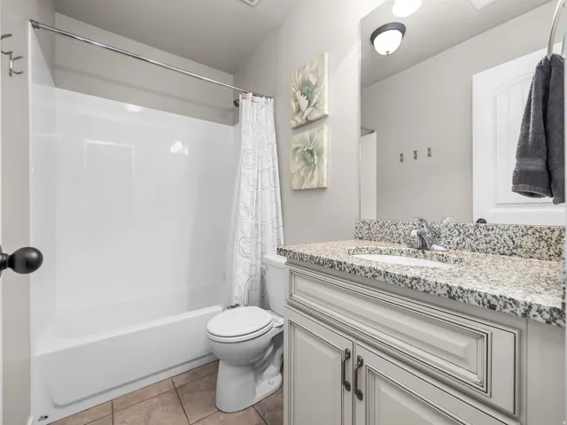 Bathroom featuring a white integrated bathtub and shower, a white toilet, a vanity with a granite countertop, a single basin sink with a chrome faucet, and tile flooring