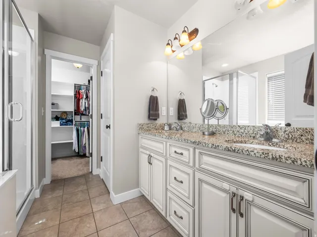 Dual vanity with granite countertop, white cabinetry with dark hardware, tile flooring, and a glass-enclosed shower
