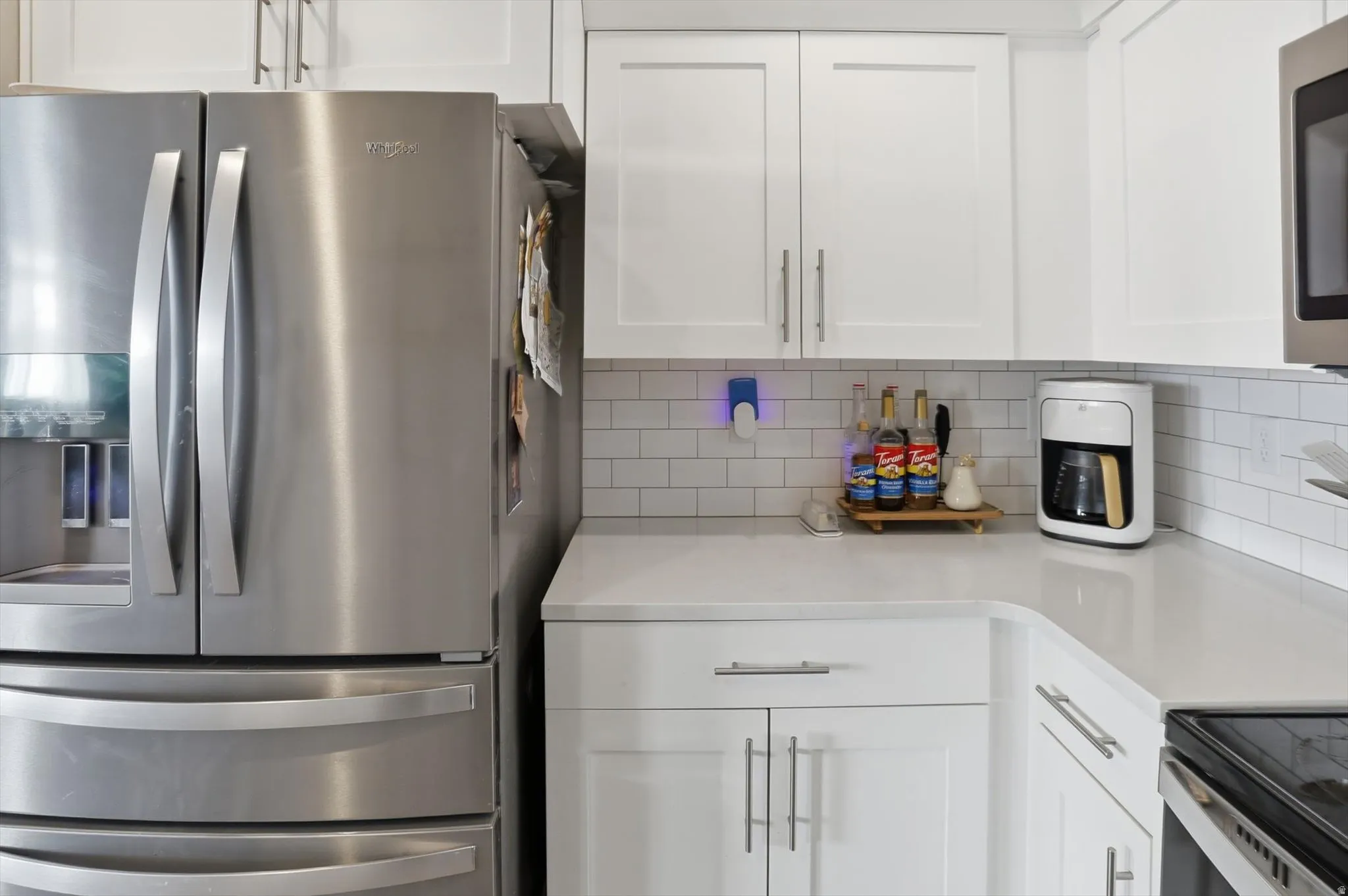 Stainless steel French door refrigerator, white shaker-style cabinetry, light-toned countertop, and subway tile backsplash