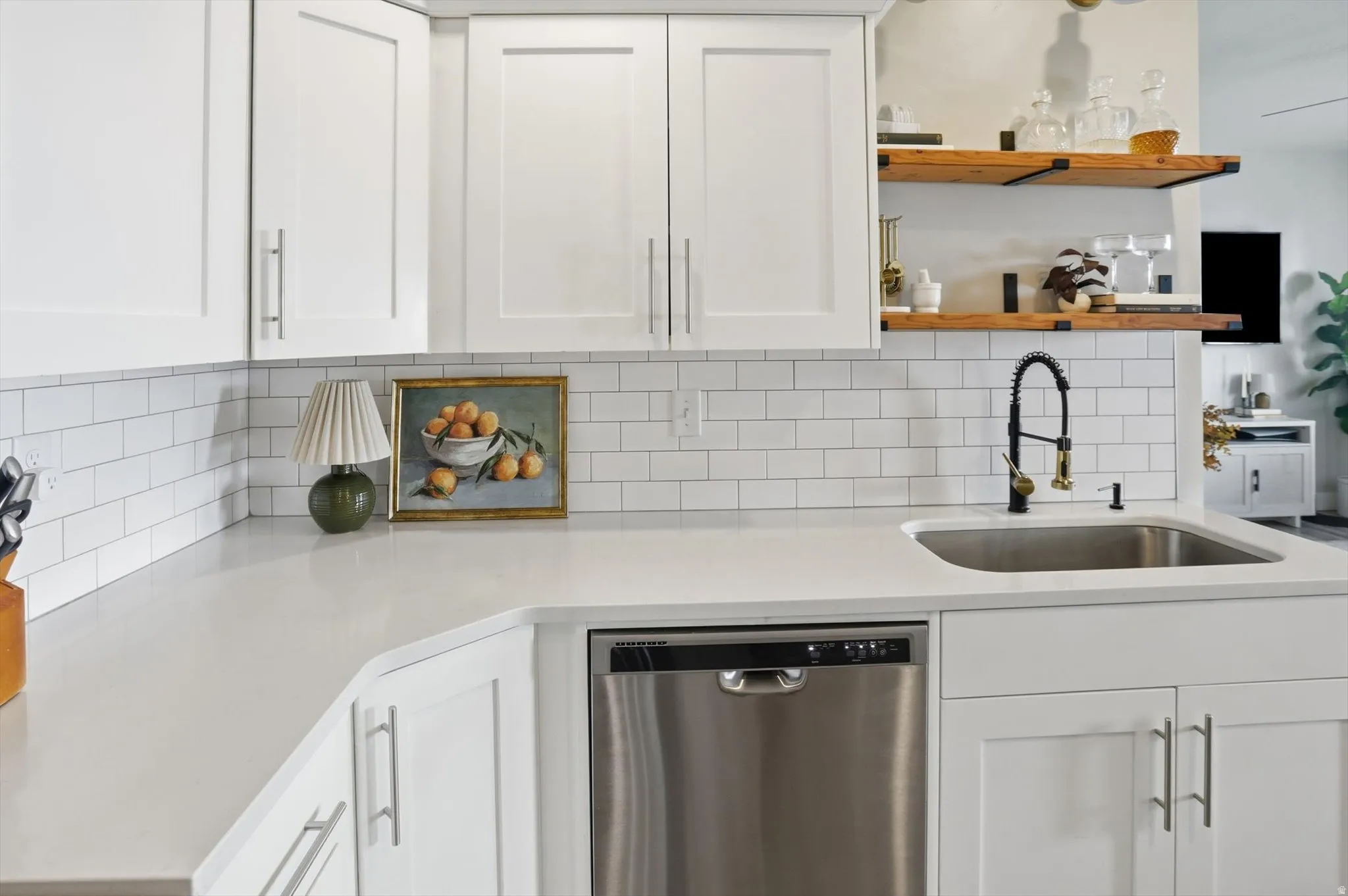 White shaker cabinetry with brushed nickel hardware, light-toned countertops, white subway tile backsplash, stainless steel sink with a matte black faucet, and open wood shelving