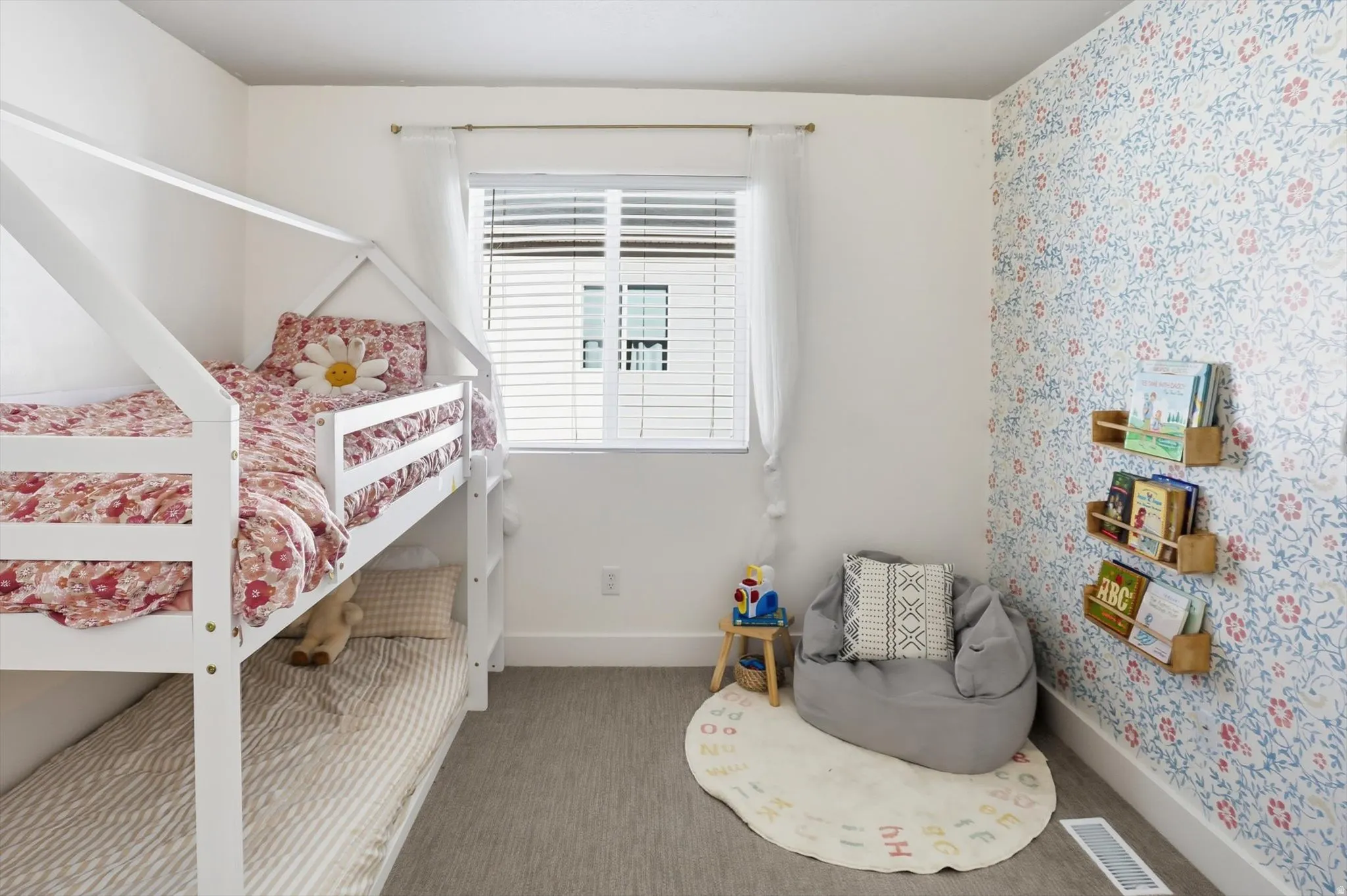 White walls with an accent wallpaper featuring a floral pattern, a window with blinds and sheer curtains, and light gray carpeting