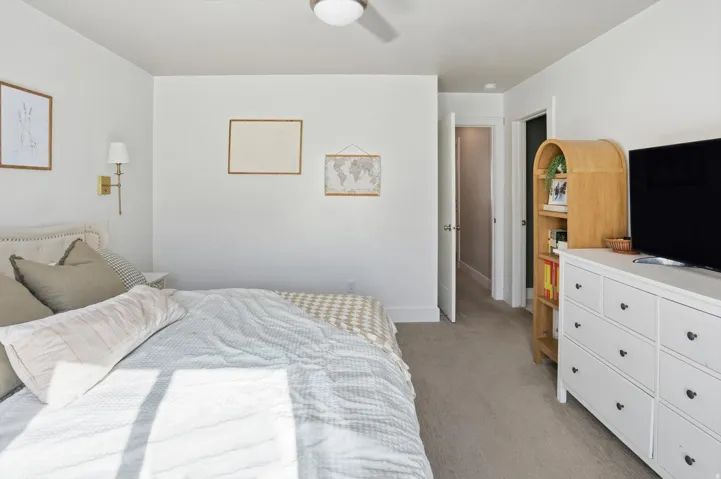 White walls, wall-mounted light fixture, light-toned carpeting, white baseboards, and a white ceiling fan