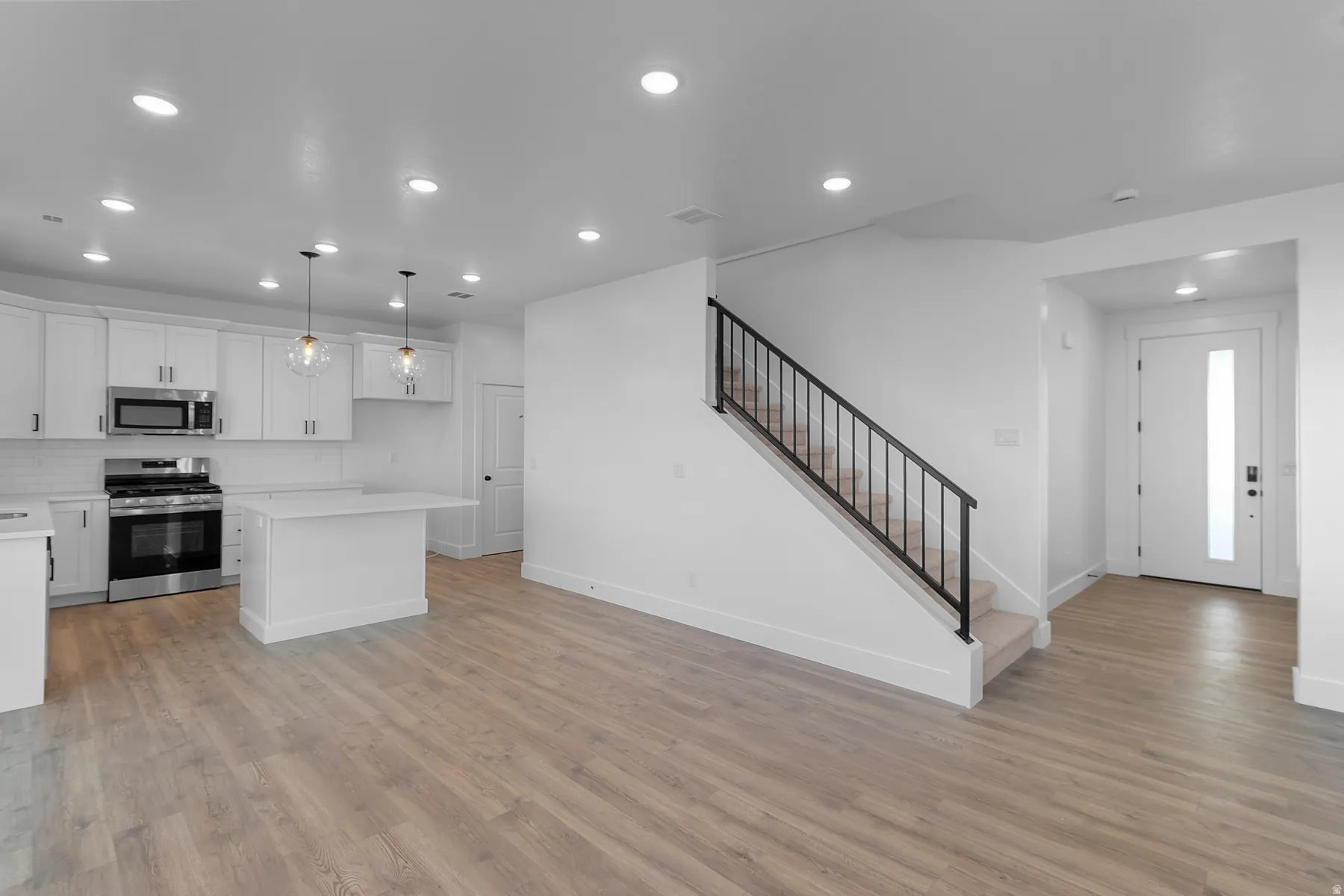 Open-concept living area featuring wood-finish flooring, white cabinetry, stainless steel appliances, and a staircase with a black metal railing