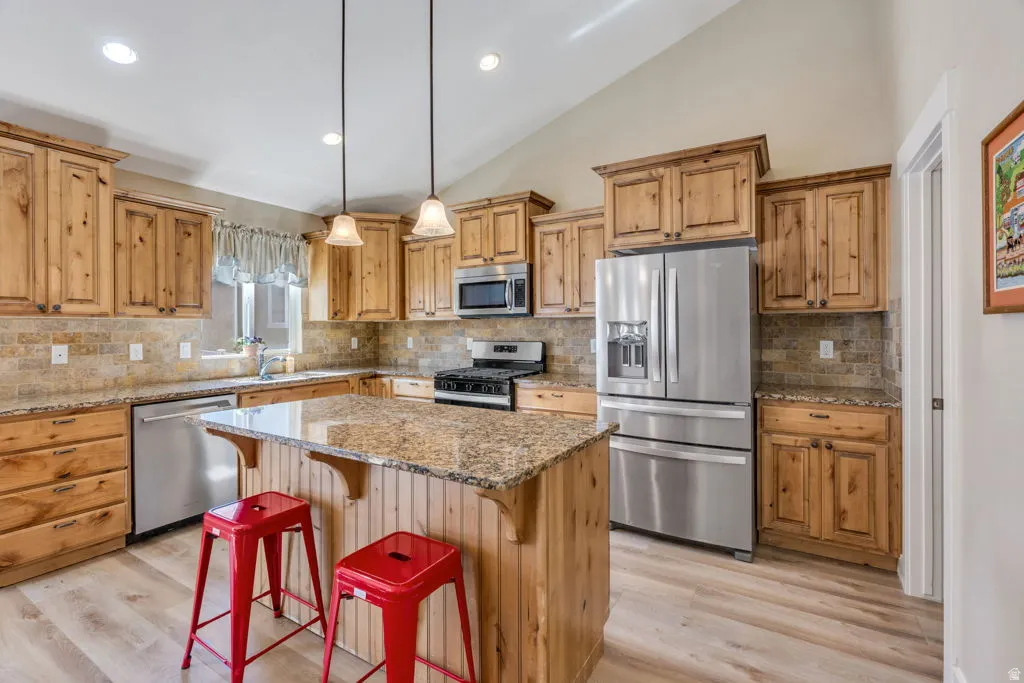 Kitchen featuring wood cabinetry, stainless steel appliances, a center island with a stone countertop, a tile backsplash, and wood-finish flooring
