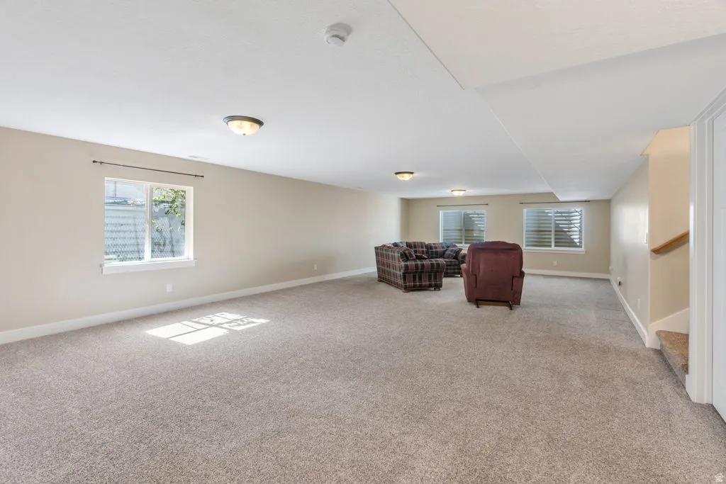 Spacious room with plush carpeting, neutral wall finishes, multiple windows, recessed ceiling lighting, and a visible staircase with a wooden handrail