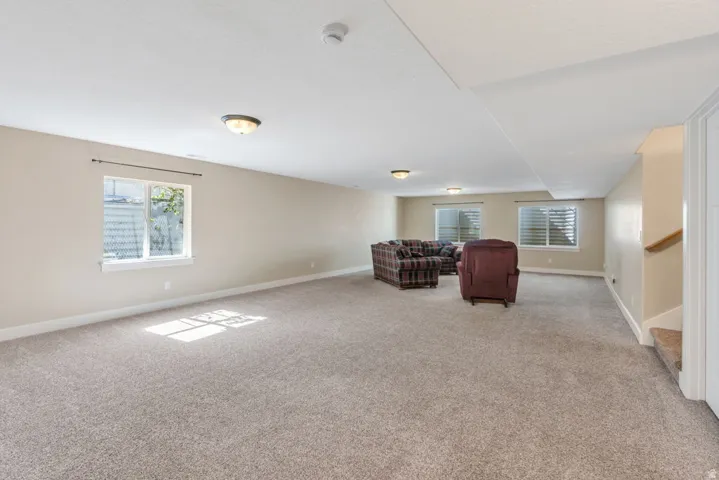 Spacious room with plush carpeting, neutral wall finishes, multiple windows, recessed ceiling lighting, and a visible staircase with a wooden handrail