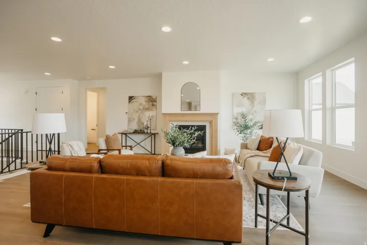 Spacious open-concept living area featuring wood-finish flooring, recessed lighting, multiple windows providing natural light, a fireplace with a wood mantel, and a black metal staircase railing