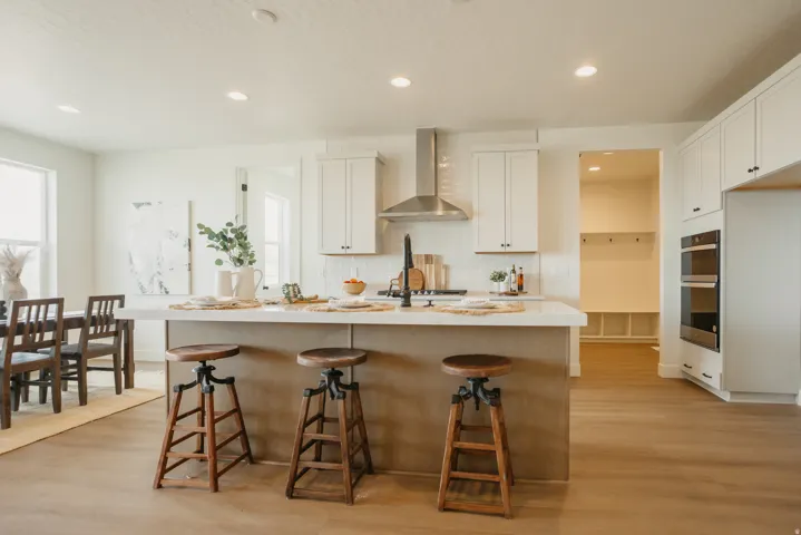 Kitchen island with counter seating, built-in double ovens, stainless steel range hood, white shaker cabinetry, and wood-finish flooring