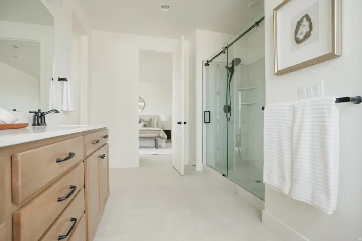 Light wood-finish vanity with dark hardware, a spacious walk-in shower with a glass enclosure, a matte black shower system, and light-toned tile flooring