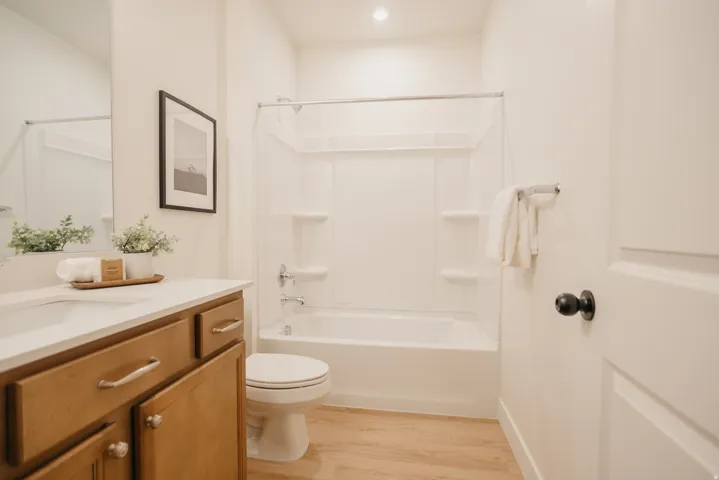 Bathroom featuring a wood-finish vanity with an integrated sink, a full-size mirror, a white toilet, a shower-tub combination, and wood-finish flooring