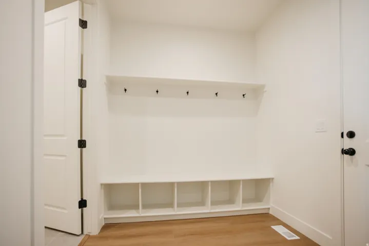 Built-in storage bench with cubbies, upper shelving with coat hooks, wood-finish flooring, and white paneled doors with matte black hardware