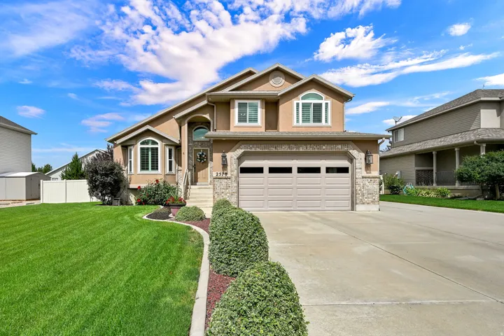 Two-story residence featuring a stucco exterior, stone facade details, a two-car garage with transom windows, a concrete driveway, and a landscaped front yard with shrubs