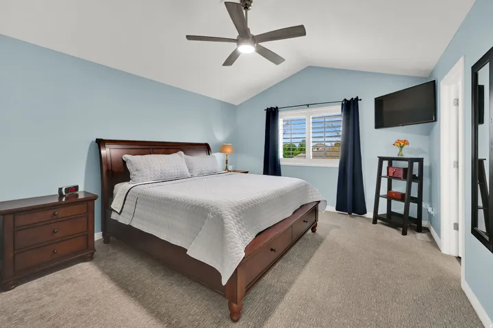 Vaulted ceiling, ceiling fan, wall-mounted television, window with plantation shutters, and light-colored carpeting