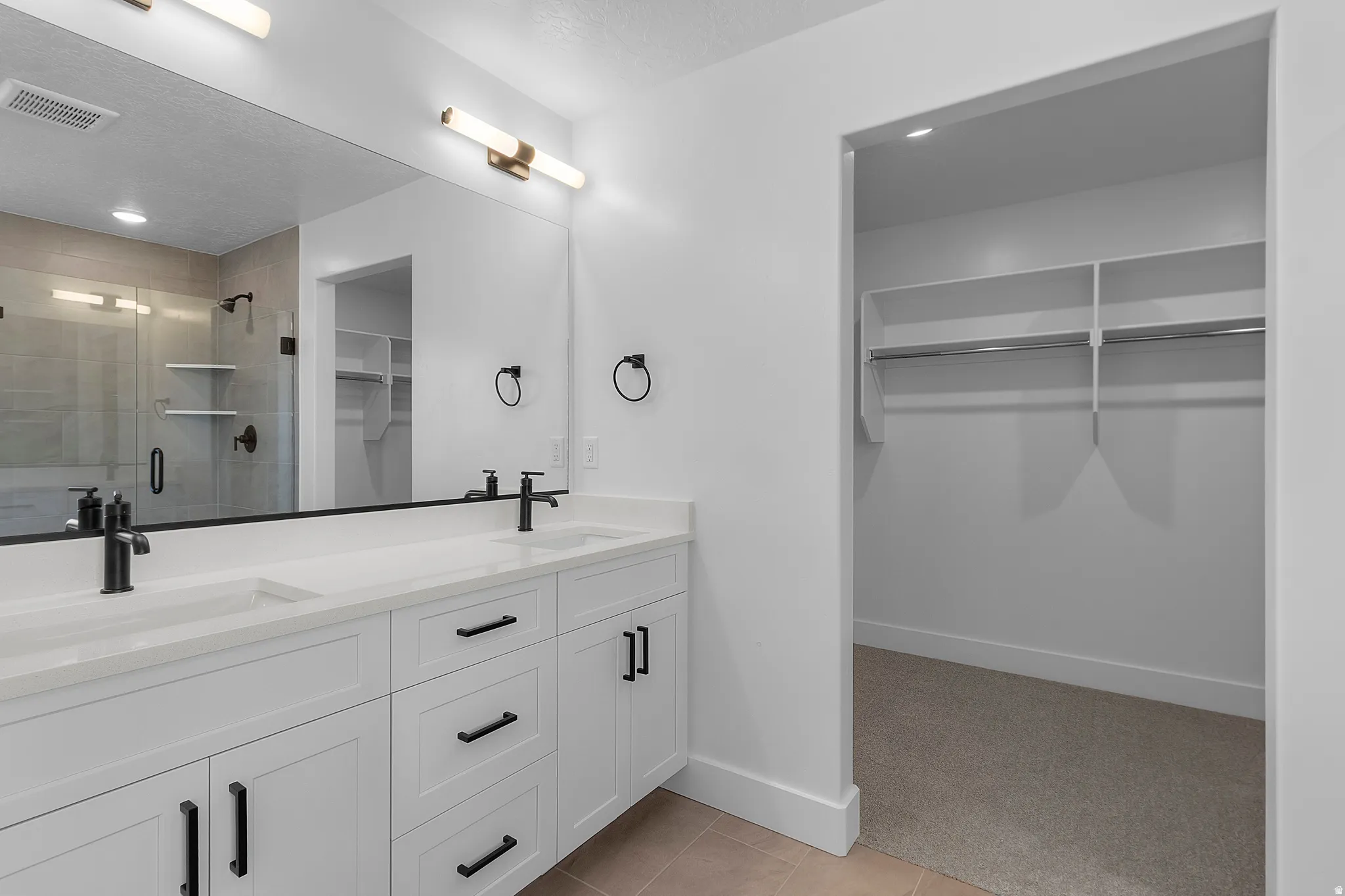 Dual sink vanity with white cabinetry, matte black fixtures, full-width mirror, and a walk-in closet with built-in shelving