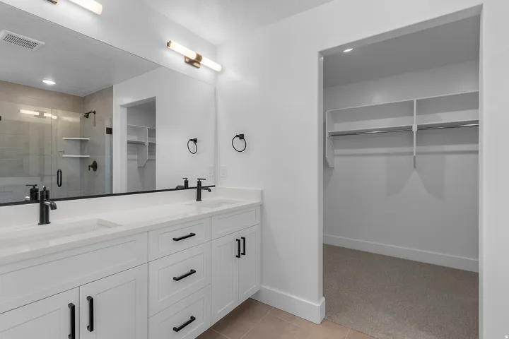 Dual sink vanity with white cabinetry, matte black fixtures, full-width mirror, and a walk-in closet with built-in shelving