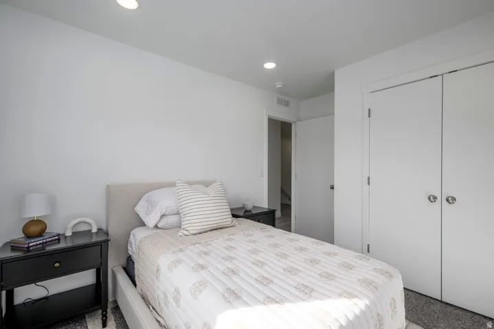 Bright secondary bedroom featuring recessed ceiling lighting, a double-door closet with white finish, carpeted flooring, and a black nightstand with open shelving