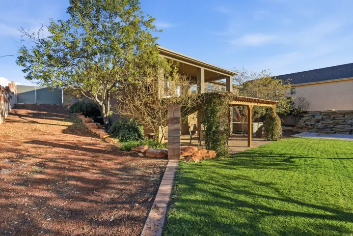 Expansive backyard featuring a lush lawn, a covered patio, a wood pergola with climbing foliage, and a stone retaining wall