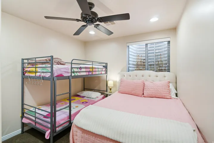 Bright secondary bedroom featuring a ceiling fan with integrated lighting, large windows with plantation shutters, metal bunk bed frame with storage shelving, and wood-finish flooring throughout