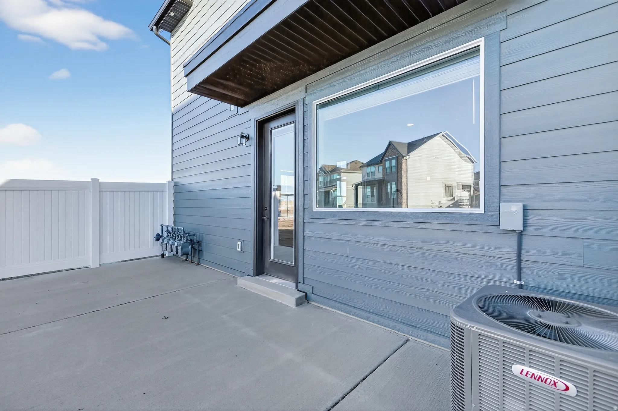 Concrete patio with a full-light glass door, large horizontal window, white privacy fence, and blue horizontal siding