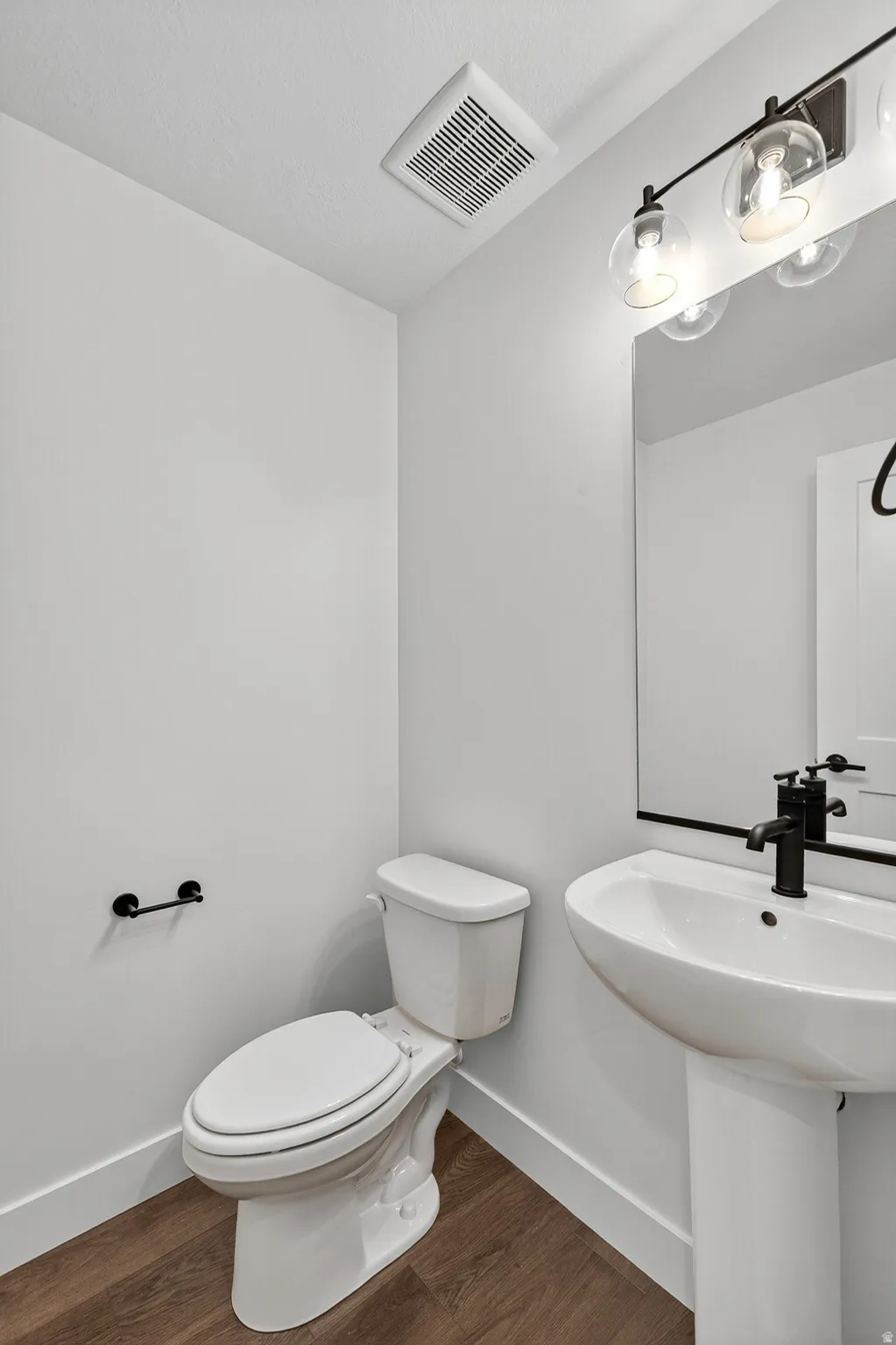 Pedestal sink with a matte black faucet, wall-mounted mirror, and wood-finish flooring